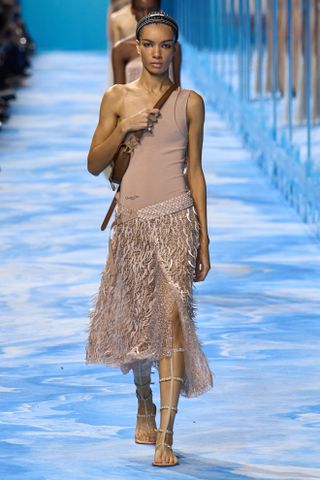 A woman wears a beige one-shoulder bodysuit, fringe, skirt, brown crossbody bag, and gold gladiator sandals.