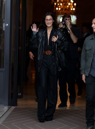 Bella Hadid in Paris for Fashion Week wearing a black '80s leather jacket, a low-cut black top, a brown belt, and pinstripe pants, all by Saint Laurent.