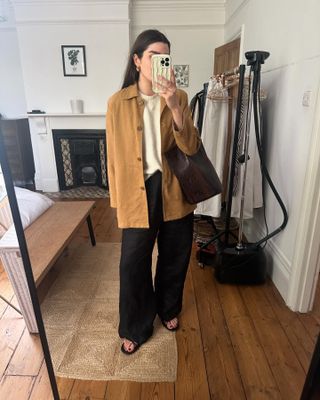 British fashion influencer Anna Howard of The Anna Edit poses for a mirror selfie wearing a camel jacket, ivory sweater, black linen pants, a burgundy snakeskin tote bag, and flat black sandals