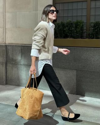 British fashion influencer Lucy Alston wearing oversize sunglasses, a beige sweater layered over an oversize white button-down shirt, black cropped pants, tan suede tote bag, gold anklet, and black Mary Jane flats