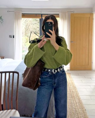 British fashion influencer Jessica Skye poses for a mirror selfie wearing a green crewneck sweater, brown suede tote bag, studded Khaite belt, and jeans