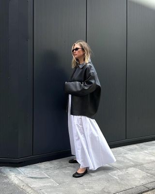 British female fashion influencer Alexis Foreman poses on a London sidewalk wearing an oversize black leather jacket, full white mixi skirt, and black Martin Margiela ballet flats