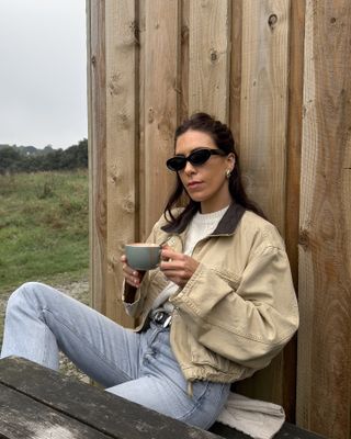 British female fashion influencer Jessica Skye sits at an outdoor cafe in Cornwall wearing black oval sunglasses, chunky silver earrings, a ruched waist barn coat, ivory cable knit sweater, black studded Khaite belt, and light-wash jeans