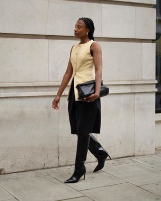 British female fashion influencer Taffy Msipa wearing a butter yellow vest top, black Bermuda shorts, black clutch, black patent knee-high pointed-toe boots