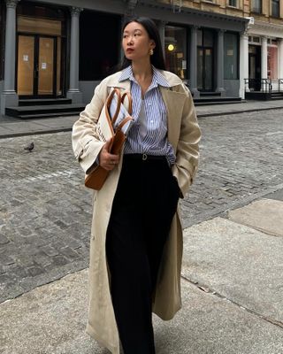 Woman wears blue striped shirt, beige trench coat, black trousers