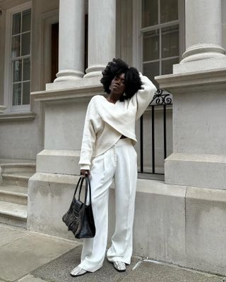 Danielle wears a white sweater, white pants, snakeskin shoes, and a black tote bag.