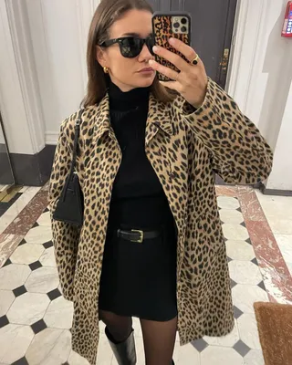 French fashion influencer Anne-Laure Mais poses for a mirror selfie wearing a leopard-print coat, black turtleneck sweater, black belted mini skirt, sheer black tights, and black knee-high boots