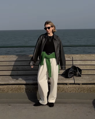 British female fashion influencer Brittany Bathgate poses next to a bench on the Kent coast wearing black sunglasses, a black leather jacket, black top, green sweater tied around the waist, belted white trousers, and black boat shoes