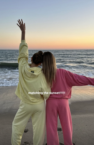 Daily Drills founders Kennedy Crichlow and Mary Ralph Lawson wearing white and pink sweatsets with back towards camera on beach.