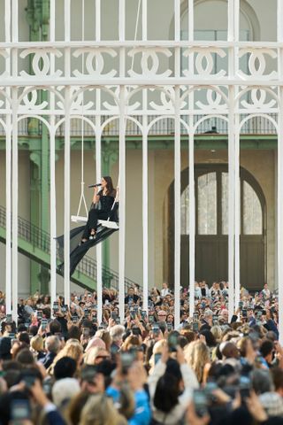 Riley Keough sitting on a swing in the Grand Palais wearing all black Chanel at the Spring Summer 2025 runway show singing 