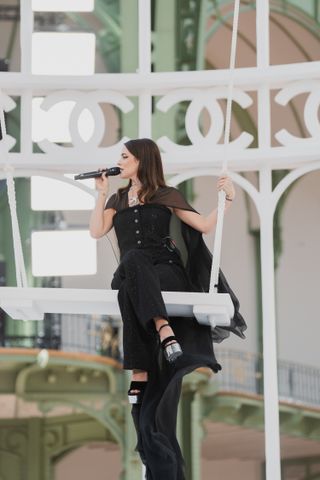 Riley Keough sitting on a swing in the Grand Palais wearing all black Chanel at the Spring Summer 2025 runway show singing 