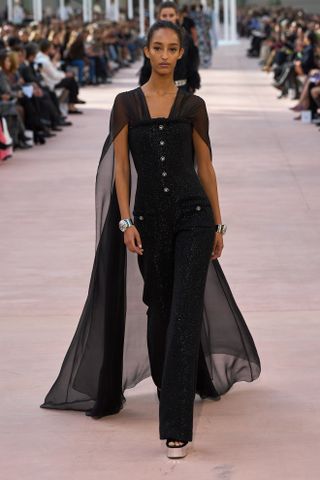 Mona Tougaard walking in the SS25 Chanel show wearing a black tweed jumpsuit with a chiffon train and platform heels.