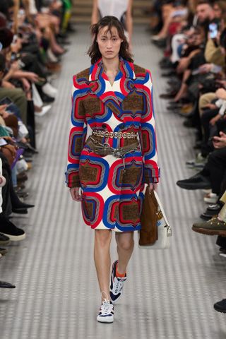 An image of a model wearing a patterned double-breasted coat with chain belts on top at the Miu Miu spring summer 2025 runway show in Paris.