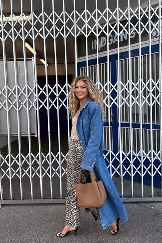 A woman walking down the sidewalk wearing a denim trench coat over a tan tank top with cheetah print jeans and black-heeled sandals.