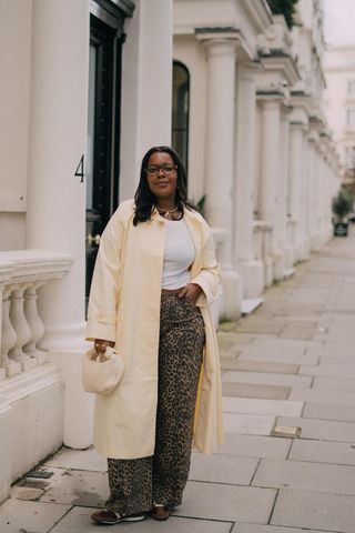 A woman walking down the sidewalk wearing a butter-yellow trench coat over a white tank top with leopard-print jeans and brown sneakers.