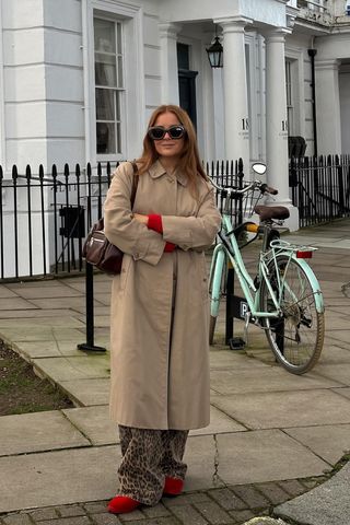 A woman standing on the street wearing a tan trench coat over a red sweater with leopard print pants and red ballet flats.