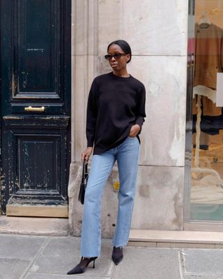 fashion influencer Sylvie Mus poses on a Paris sidewalk wearing oval sunglasses, a black crewneck sweater, mini black bag, straight-leg raw-hem jeans, black tights, and black pumps