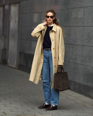 fashion influencer Anouk Yve stands on a sidewalk wearing oval sunglasses, collared trench coat, black sweater layered over a white tee, brown suede tote bag, a black belt, straight-leg jeans, white socks and loafers