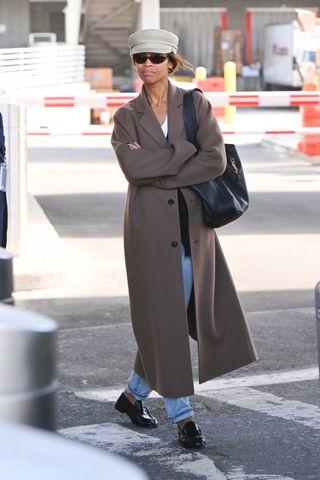 Zoe Saldana is seen at John F. Kennedy International Airport on October 14, 2024 in New York City wearing a hat, brown coat, black cardigan, jeans, and black Saint Laurent loafers. She is carrying a Saint Laurent shoulder bag.