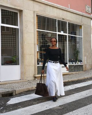 Taffy wears a black boatneck long-sleeve top, black stud belt, white maxi skirt, black shoes, and a brown suede tote bag.