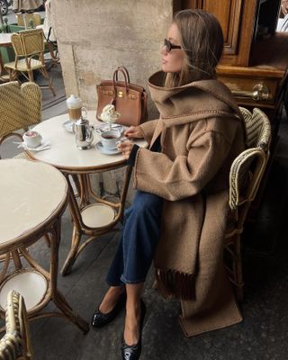 Lovisa wears a tan scarf coat, blue jeans, black ballet flats, and a tan Hermes bag.