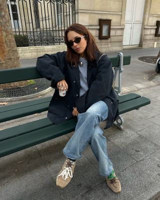 Debora wears a navy barn coat, gray sweater, blue jeans, and brown suede Miu Miu sneakers.