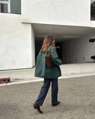 Jen wears a green barn coat, brown bag, blue jeans, and brown suede boots.