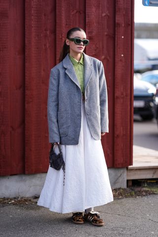 A woman wears a grey blazer, white maxi skirt and leopard print trainers whilst standing in front of a red building