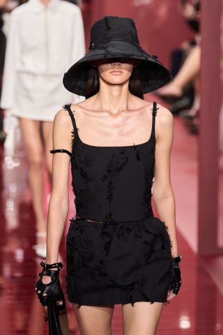 Model wearing a black sun hat with a black sleeveless top and miniskirt at the Gucci S/S 25 runway show.