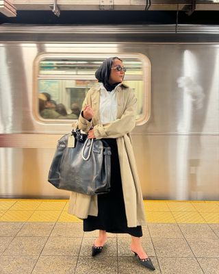 Woman wearing white button-down, black skirt, Coach bag and trench coat while standing in front of New York City subway.