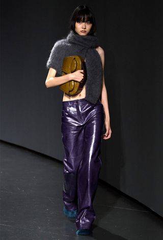 Model walking the 16Arlington runway show wearing a gray tied sweater with purple leather pants, a green handbag, and blue flats.