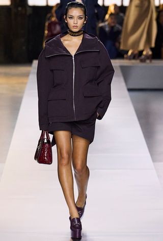 Model walking the Gucci fall runway show wearing a purple jacket with a miniskirt, platform loafers, and a red bag.