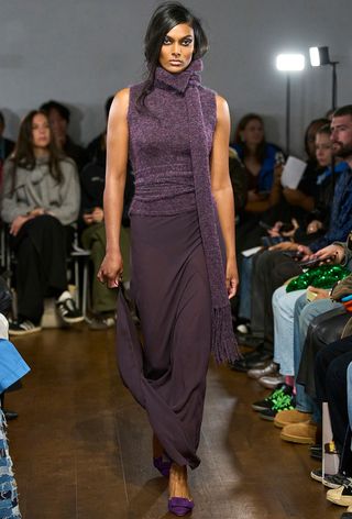 Model walking the Aaron Esh runway show wearing a purple sweater with a built-in scarf, maxi knit skirt, and pointed pumps.