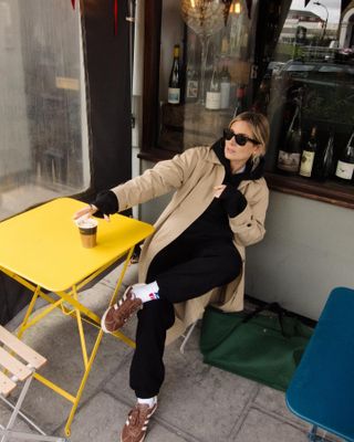 Lucy Williams wearing a black sweatsuit and a classic trench coat with brown Adidas sneakers at a coffee shop in London.