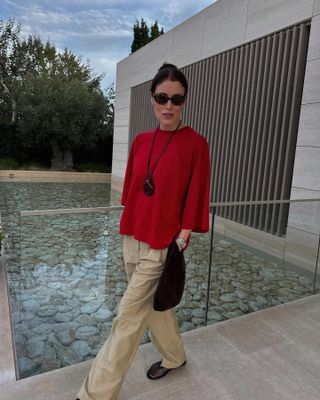 Annabel wears a red top, long necklace, black bag, tan trousers, and black mesh flats.