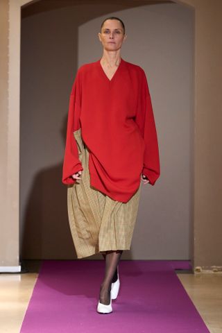 Adam Leenaerdt F24 model wears an oversize red sweater, baggy skirt, sheer tights, and white pumps