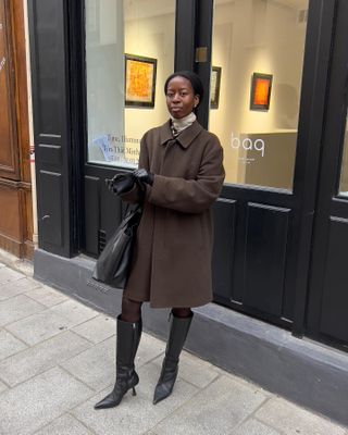 Woman wearing brown coat, black boots, and black bag while standing on Paris street.