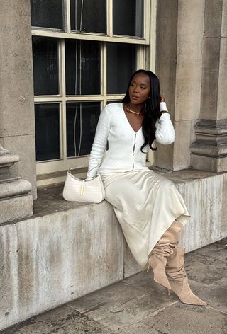 best suede boots shown on a photo of a woman sitting outside on a window ledge wearing a white cardigan with a white slip skirt and light tan slouchy suede boots