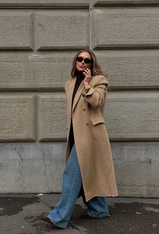 best suede boots shown on a photo of a woman walking down the street wearing a tan long coat over a black turtleneck, wide-leg jeans, and brown pointed ankle boots