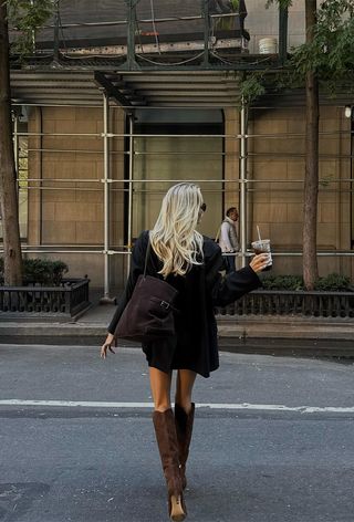 best suede boots shown on a photo of a woman walking in the street in New York wearing an oversize black blazer over a black dress with a large dark brown suede bag and knee-high suede brown boots