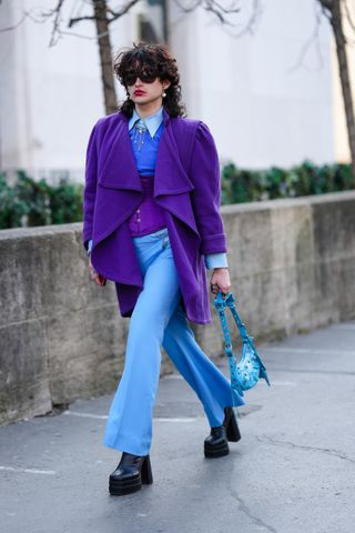 A guest wears black sunglasses, a pale blue shirt, a royal blue pullover, a sugilite purple corset, a neon purple wool coat, pale blue flared pants, during Paris Fashion Week - Womenswear Fall Winter 2023 2024, on March 01, 2023 in Paris, France.