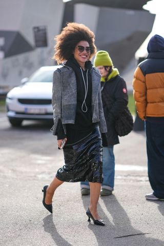 A woman wears a black sequin skirt with a black sweatshirt and gray jacket