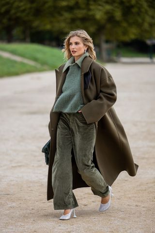 A woman wears a sage green cashmere collared sweater with olive green pants and a brown coat.