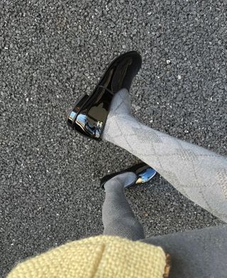 A photo of a woman wearing gray knit tights, a yellow tweed jacket, and black patent leather Chanel moccasins.