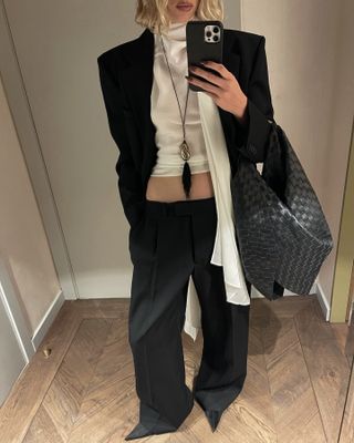 Elsa wears a black blazer, white silk top, black bottega bag, black trousers, and satin heels.