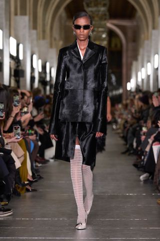 Model wearing a black calf hair blazer and skirt with white thigh-high socks at the Tory Burch F/W 24 show.