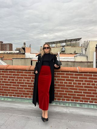 Eliza Huber wearing a black boatneck T-shirt with a black long coat and red velvet Blazé Milano midi skirt.