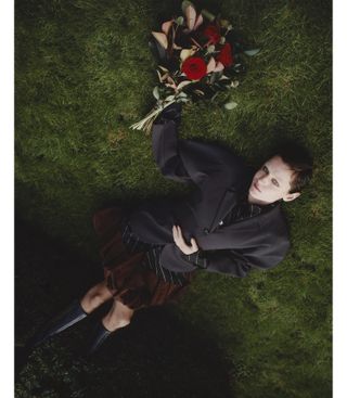 Emma Corrin lays in the grass holding a bouquet of red roses wearing an oversize Renaissance Renaissance blazer and skirt.