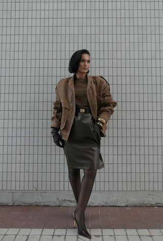 one of the best chunky cuff bracelets is shown in a photo of a woman standing outside wearing a brown leather bomber jacket over a brown blouse styled with an olive green pencil skirt, brown tights, brown slingback pumps, and brown leather gloves with chunky gold bangles layered on top of them