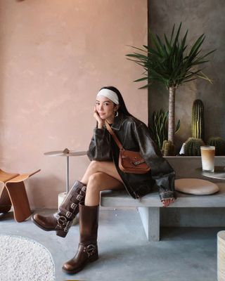 A woman is seated wearing a brown moto jacket wearing matching knee-high boots with silver buckles.